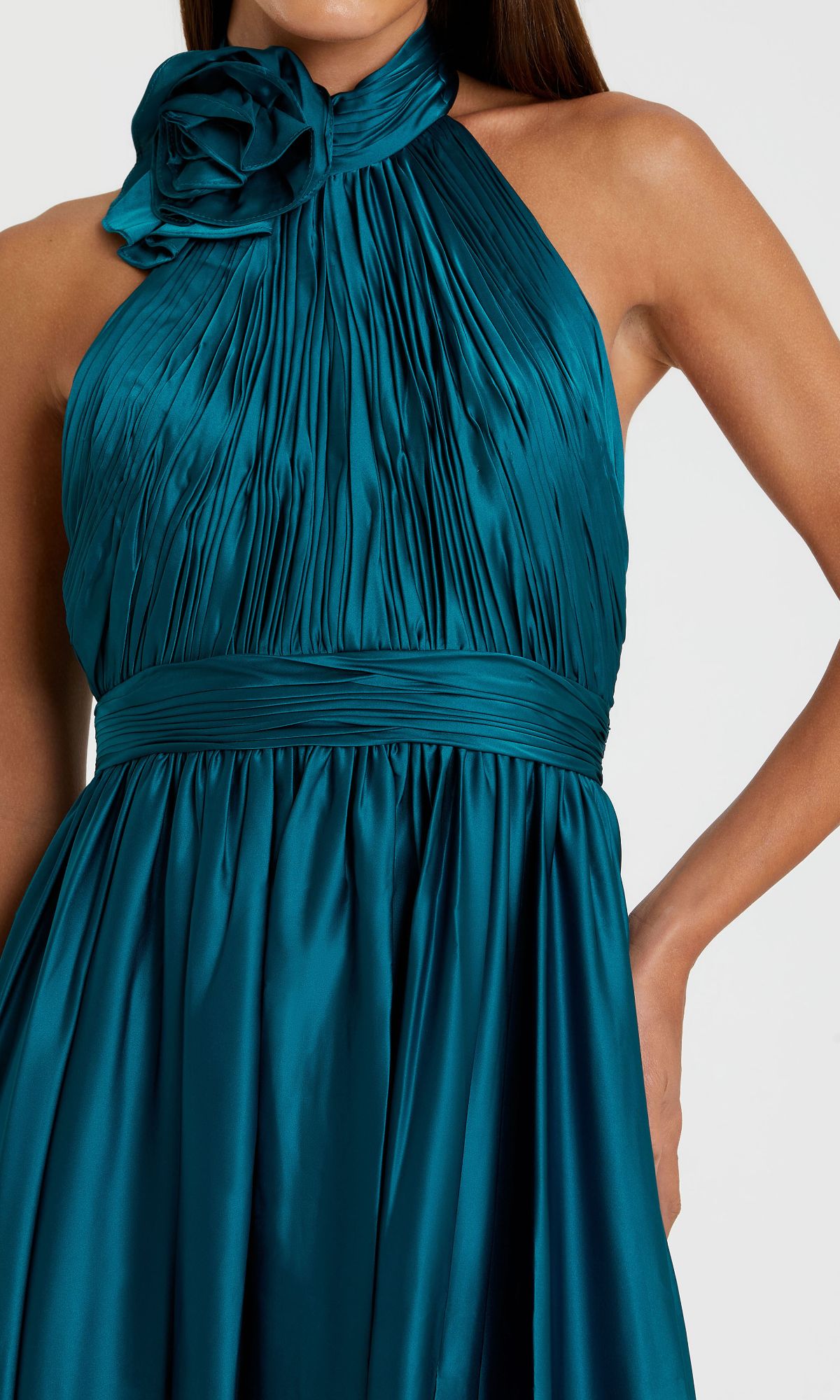 A woman wears the Halter Midi Wedding Guest Dress: Mac Duggal 49674—an ocean blue, sleeveless, pleated satin dress with a fitted waistband and large neckline flower. A chic semi-formal choice for special occasions.